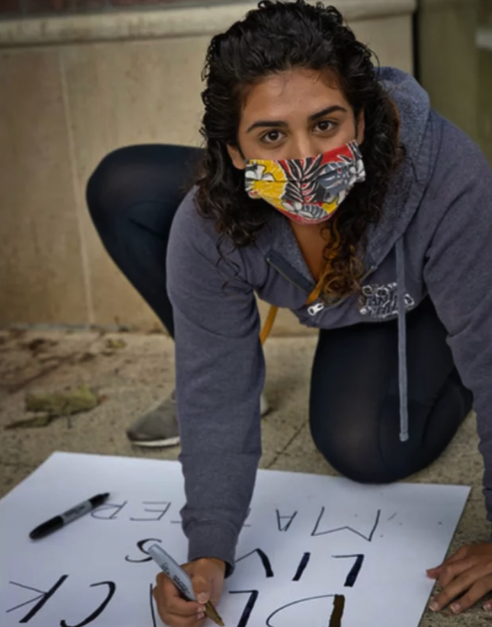 Ariana Jones, wearing a fabric mask due to the COVID-19 pandemic, leans over a poster board and creates a protest sign.